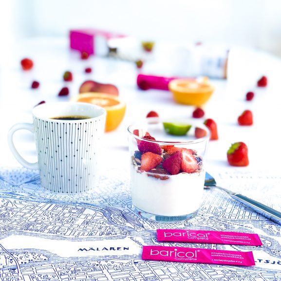 Frukostunderlägg med stockholmskarta på den står kaffekopp, glas med yoghurt med granola och jordgubbar omgiven av två dospåsar baricol pulver