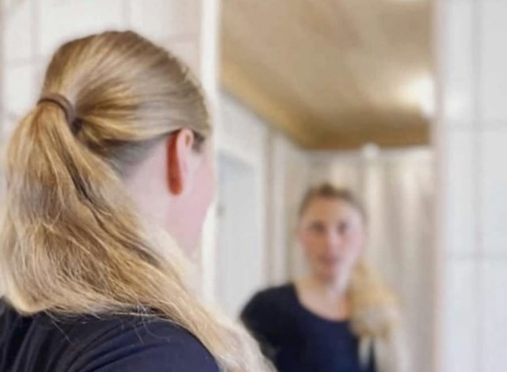Blonde woman with ponytail with body dysmorphia looking at reflection in mirror